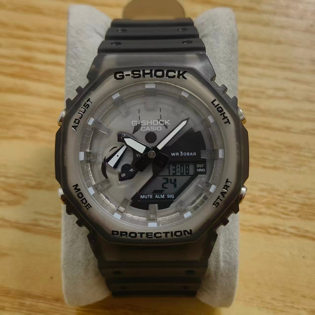 A Casio G-Shock analog-digital watch with a translucent smoky gray and black resin case, displayed on a light gray cushion over a light wood surface.
