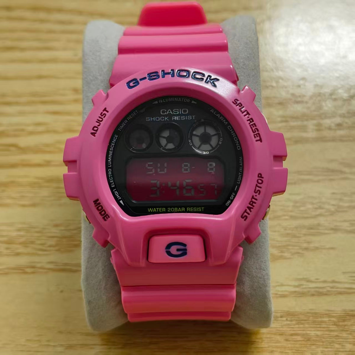 A pink Casio G-Shock digital watch with a black face, resting on a gray stand over a light wood surface.