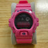 A pink Casio G-Shock digital watch with a black face, resting on a gray stand over a light wood surface.