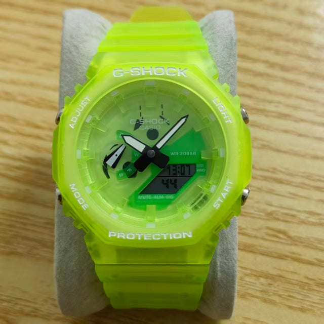 A bright neon yellow-green Casio G-Shock watch displayed on a gray stand over a light wood surface. The watch features a translucent lime green strap and case with a matching dial, black hands, and a digital display window.