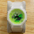 A Casio G-Shock watch with a translucent white resin case and a bright neon green dial, displayed on a gray velvet watch stand over a light wood surface.
