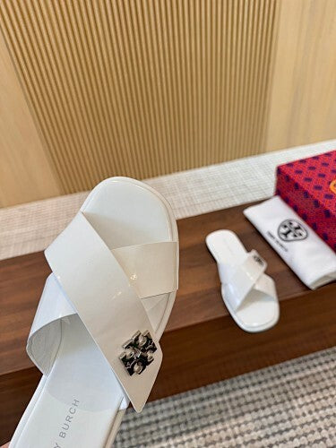 A close-up of a white Mary Burchi slide sandal with a black and white floral emblem on the strap, resting on a wooden surface next to its red and black branded box and a white dust bag.