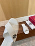A close-up of a white Mary Burchi slide sandal with a black and white floral emblem on the strap, resting on a wooden surface next to its red and black branded box and a white dust bag.