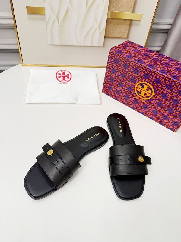 A pair of black Tory Burch slide sandals with gold-tone hardware, displayed on a white surface alongside a white dust bag with a red logo and a purple patterned box with a gold logo.