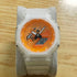 A Casio G-Shock analog watch with a translucent white resin case and strap, featuring a vibrant orange dial with black hands and a digital display, resting on a gray display stand over a light wood surface.