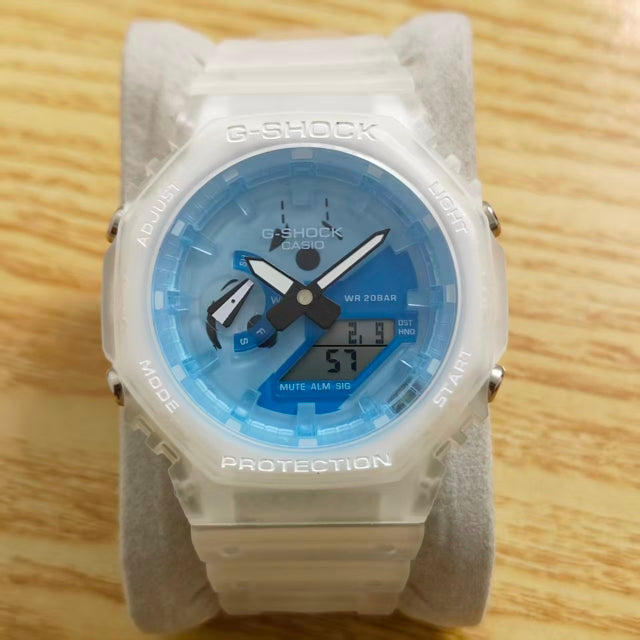 A Casio G-Shock analog watch with a translucent white case and strap, featuring a light blue and blue dial with black hands and a digital display, resting on a gray display stand over a light wood surface.