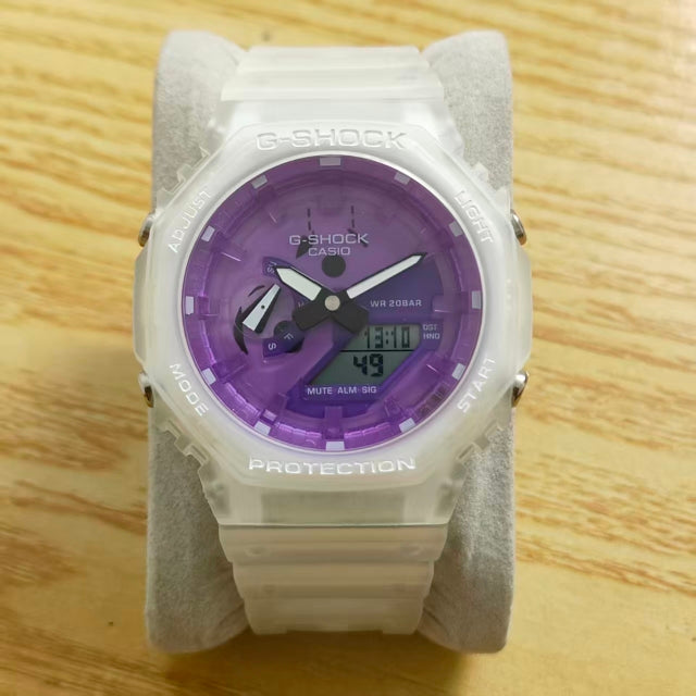 A Casio G-Shock analog watch with a translucent white case and strap, featuring a vibrant purple dial, displayed on a gray stand over a light wood background.